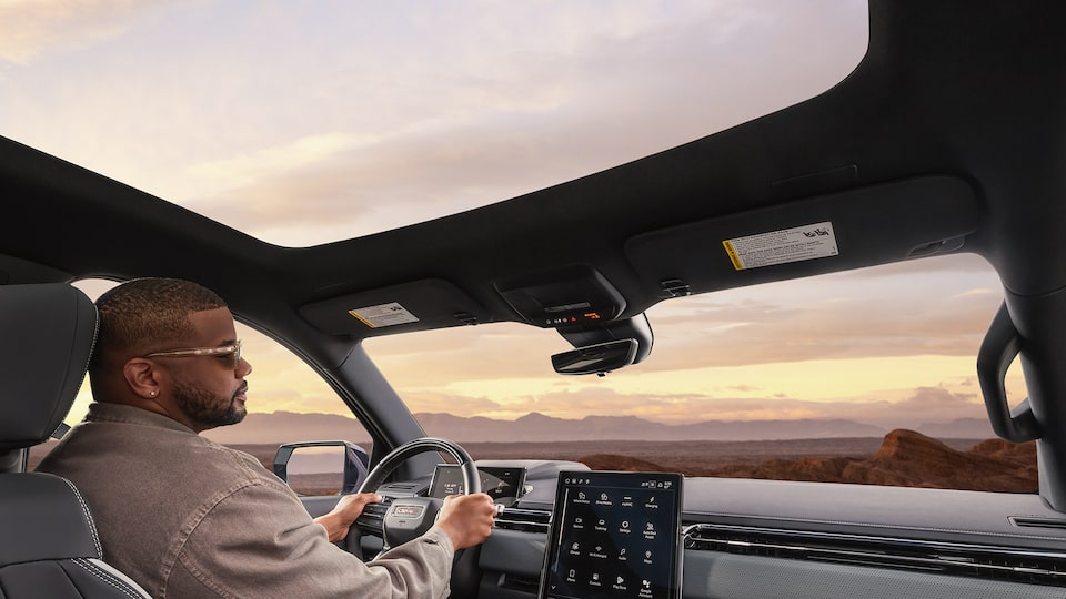 Back Seat View of a Man Driving the 2026 GMC Sierra EV with the Panoramic Sunroof Open During a Beautiful Sunset