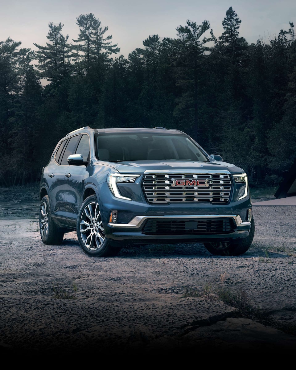 Front Three-Quarters View of a 2024 GMC Mid-Size SUV Driving on a Dirt Path with Mountains in the Background