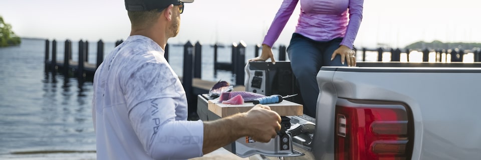 A Woman Sitting in the Truck Bed of A GMC Parked at the Pier as a Man Cleans His Freshly Caught Fish