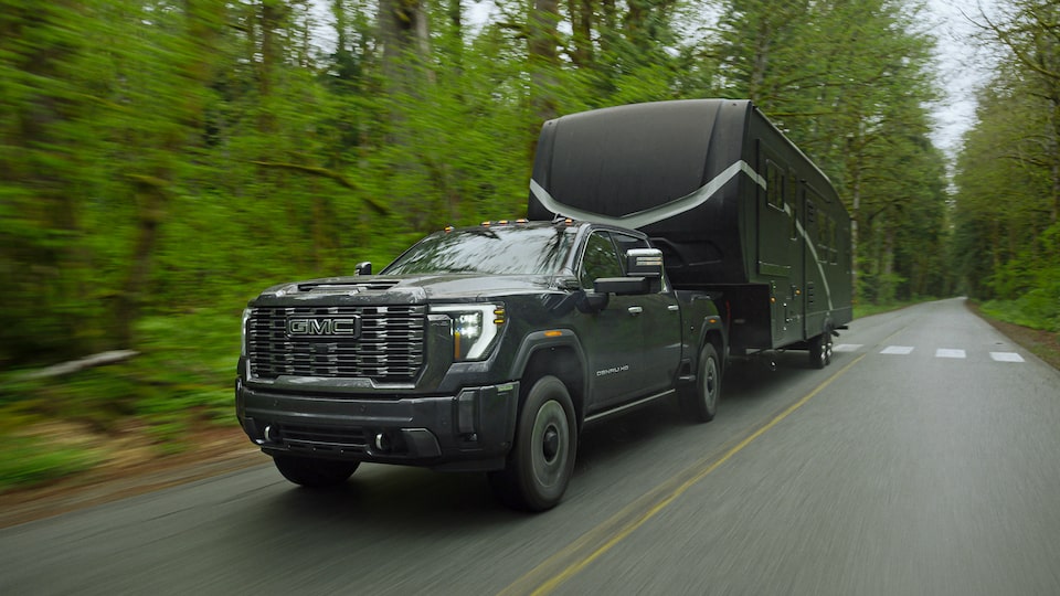 Close-Up Front Three-Quarters View of a GMC Sierra HD Denali Truck Hauling a Trailer on a Tree-Lined Road