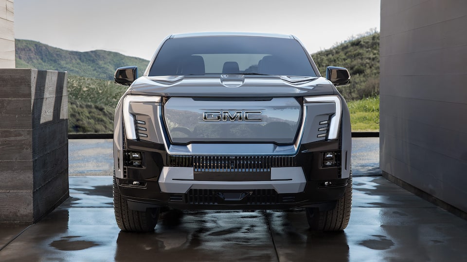 Front Three-Quarter View of the 2025 Sierra EV Pickup Truck Parked in a Driveway with the Vehicle's Headlights on